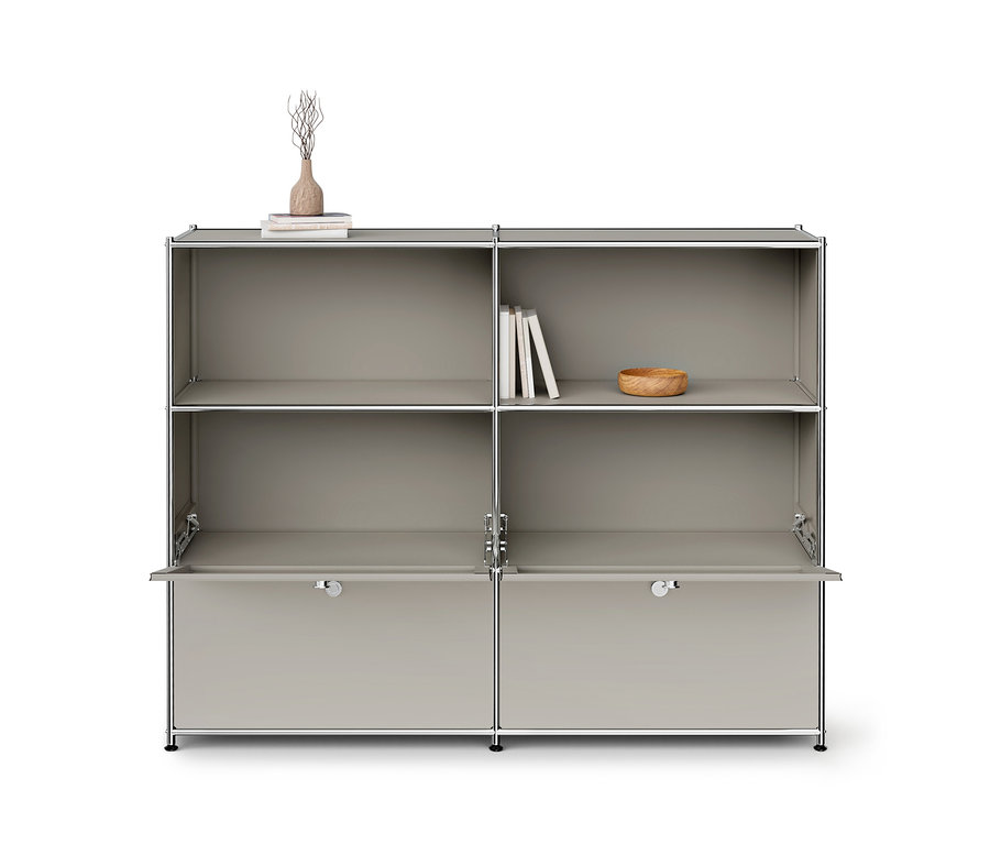 Helles graues Modulschrank mit Chromrahmen und sechs Fächern. Die oberen zwei Reihen haben offene Regale, die untere Reihe hat zwei teilweise geöffnete Klapptüren. Eine Vase mit Ästen und Bücher stehen auf dem oberen linken Regal, Bücher und eine Holzschale auf dem oberen rechten Regal.