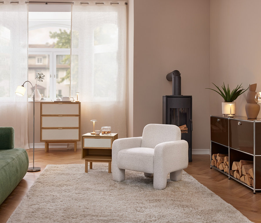 Ein gemütliches Wohnzimmer mit grünem Sofa, hellem Sessel und schwarzem Holzofen. Eine Kommode und ein Beistelltisch mit einer Trio LED-Tischleuchte »Flynt« stehen am Fenster. Rechts ein braunes Regal mit Holzscheiten und einer weiteren Tischleuchte.