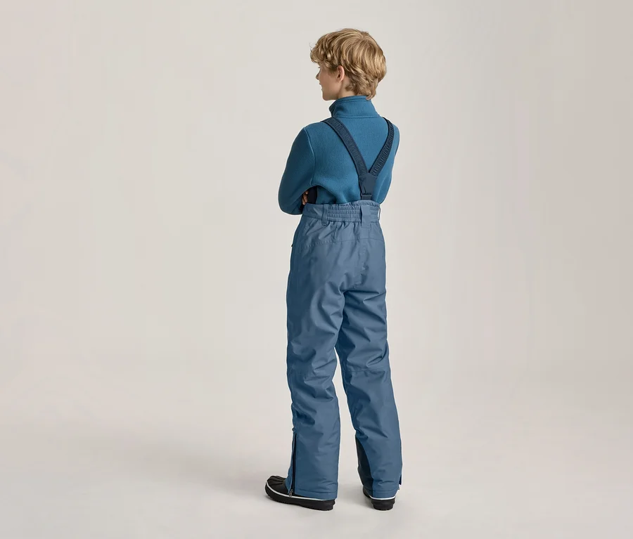 Ein junger Mensch mit kurzen, hellen Haaren steht mit dem Rücken zum Betrachter, trägt einen blauen Rollkragenpullover und eine blaue Schneehose mit Hosenträgern. Die Arme sind verschränkt.