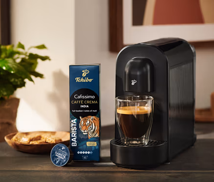Schwarze Tchibo Cafissimo Kaffeemaschine mit einem Glas Kaffee, daneben eine Packung Tchibo Cafissimo Caffè Crema India Kaffeekapseln und eine einzelne Kapsel.