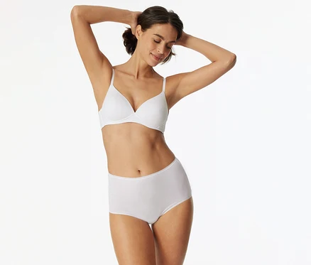 Frau mit dunklem Haar, die einen weißen BH und einen weißen High-Waist-Slip trägt, die Arme hinter dem Kopf verschränkt und lächelnd die Augen geschlossen hat.