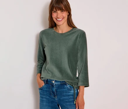 Lachende Frau trägt ein grünes CECIL 3/4-Cordshirt mit Kordelzug und blaue Jeans, eine Hand in der Tasche.