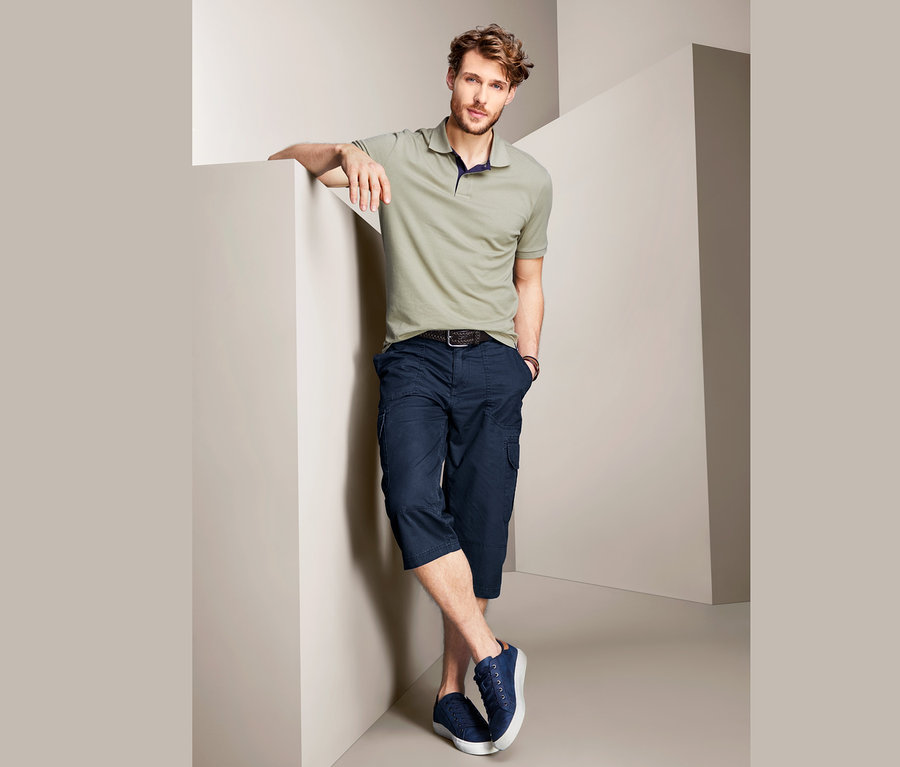 Mann an Wand angelehnt trägt olivfarbenes Poloshirt und marineblaue Cargo-Bermudas in 3/4-Länge.