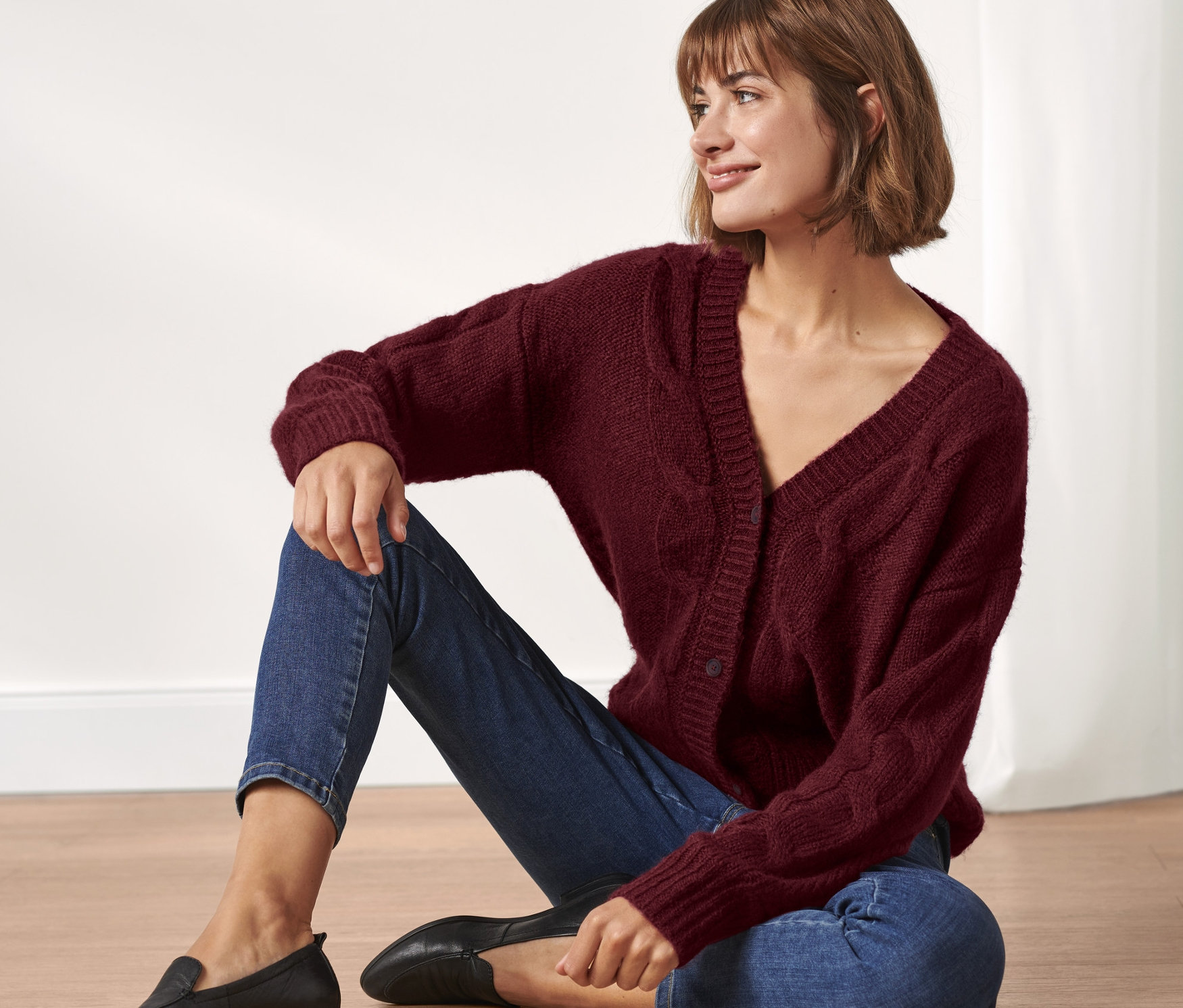 Frau sitzt auf dem Boden und trägt eine Strickjacke mit Zopfmuster, Jeans und schwarze Slipper.