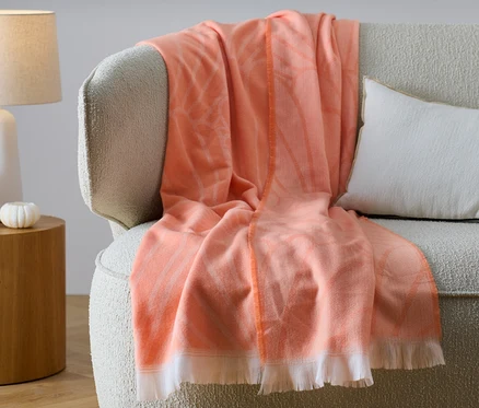 Eine orangefarbene Wende-Wohndecke mit Fransen liegt auf einem beigen Sofa neben einem weißen Kissen.