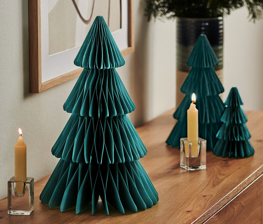 Auf einer Kommode stehen ein großer faltbarer Papierbaum, zwei kleinere Bäume, eine brennende Kerze in einem Glasleuchter und eine Vase mit Zweigen.