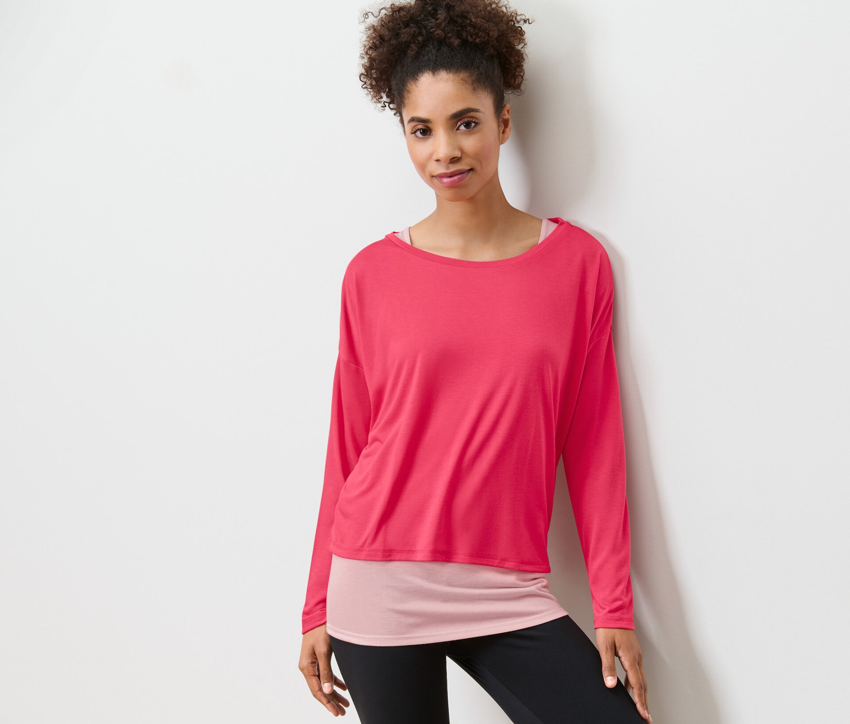 Frau posiert in einem pinken 2-in-1-Sportshirt und schwarzen Leggings.