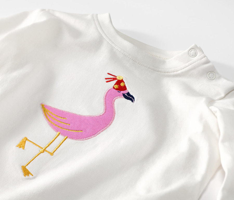 Detailansicht eines weißen Kinder-Langarmshirts mit einer aufgestickten Flamingo-Applikation mit Geburtstagshut.