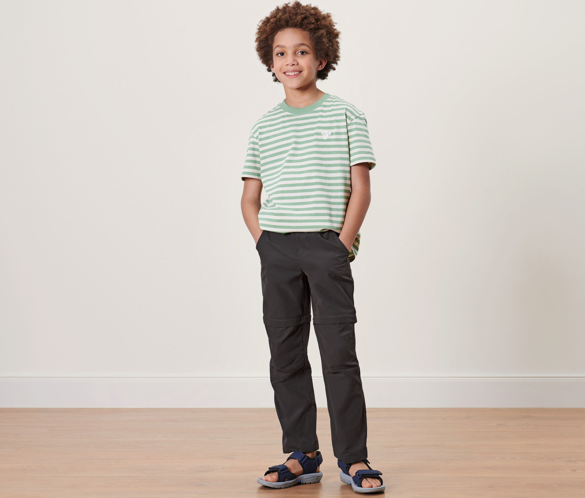 Ein Junge steht und trägt eine Kinder-Funktionshose, ein gestreiftes T-Shirt und Sandalen.