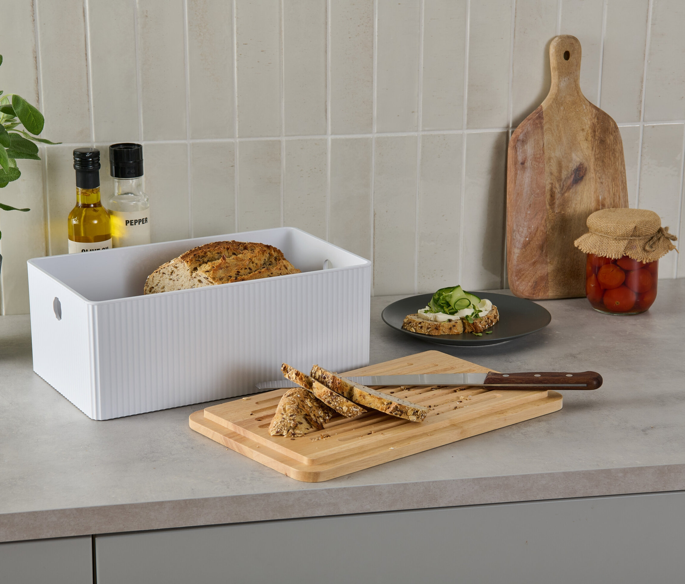 Brotkasten mit Schneidebrett auf einer Küchentheke neben Brot, einem belegten Brot, Öl, Pfeffer, einer Pflanze und eingelegten Tomaten.