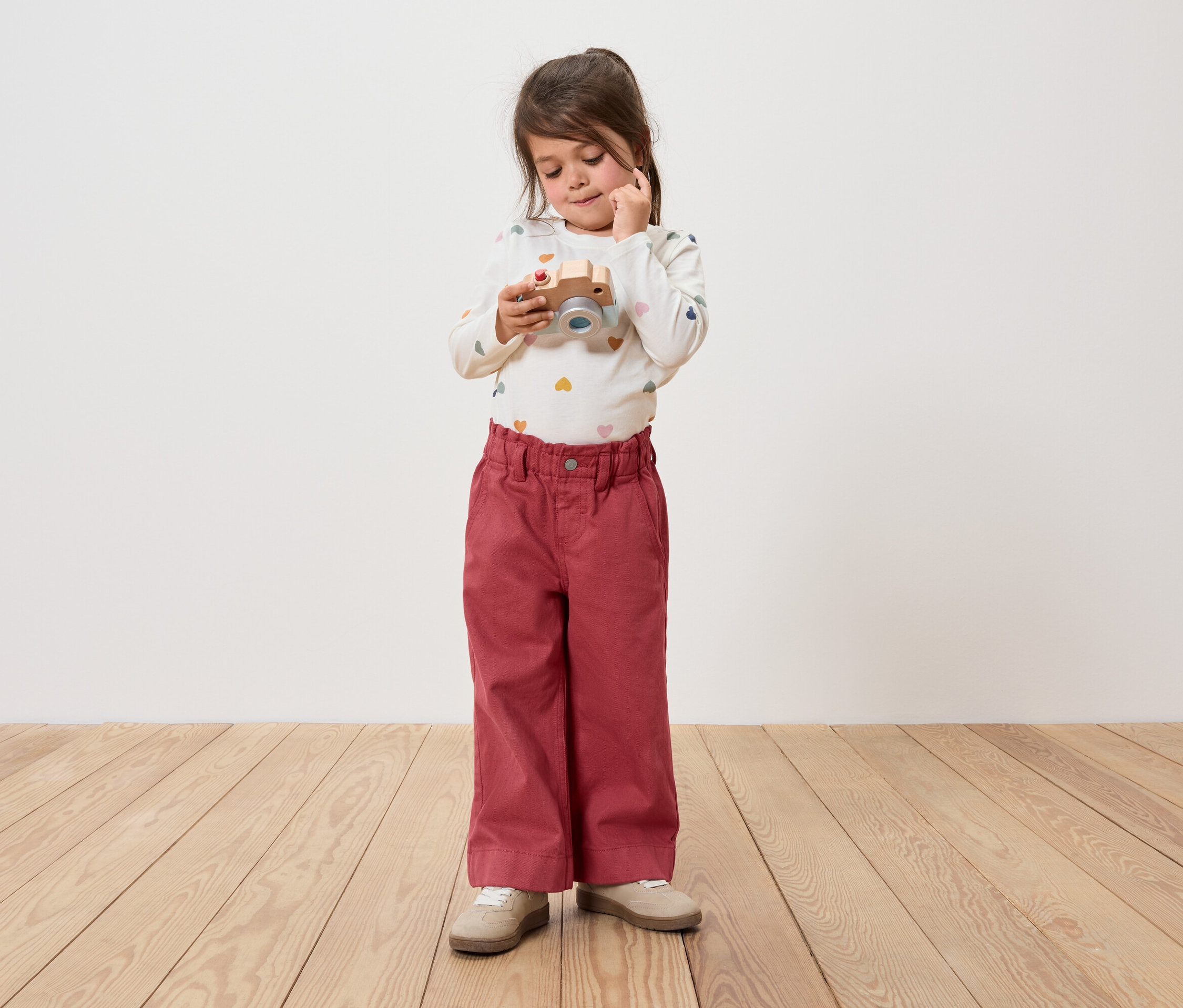 Mädchen hält eine Holzkamera und schaut sie an. Sie trägt eine Hose und ein langärmeliges Shirt.