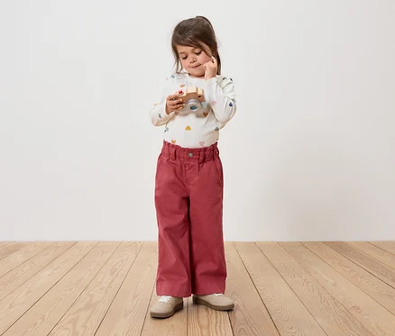 Mädchen hält eine Holzkamera und schaut sie an. Sie trägt eine Hose und ein langärmeliges Shirt.