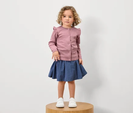 Mädchen mit lockigem Haar steht auf einem Holzsockel und trägt eine rosa Rüschenstrickjacke, einen blauen Rock und weiße Turnschuhe.