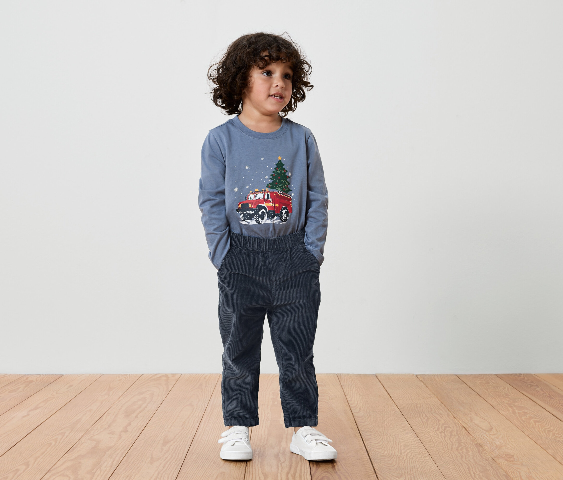Junge mit braunen Haaren trägt ein blaues T-Shirt mit einem roten Auto und einem Weihnachtsbaum darauf, dazu dunkelblaue Hosen und weiße Turnschuhe.