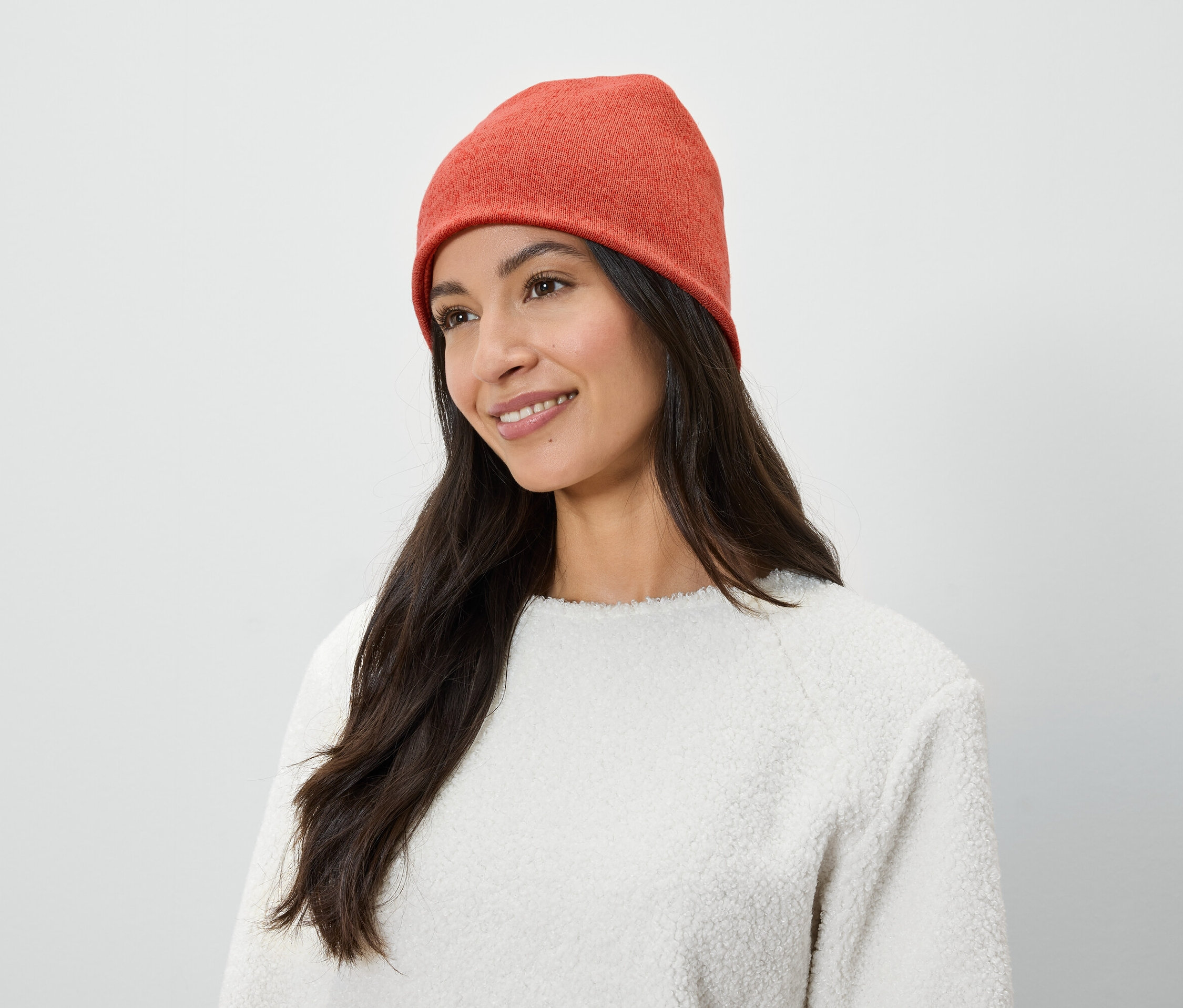 Frau mit langen braunen Haaren und einem Lächeln trägt eine orangefarbene Mütze und einen weißen Pullover.