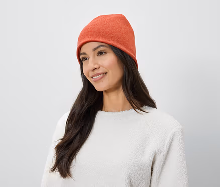 Frau mit langen braunen Haaren und einem Lächeln trägt eine orangefarbene Mütze und einen weißen Pullover.