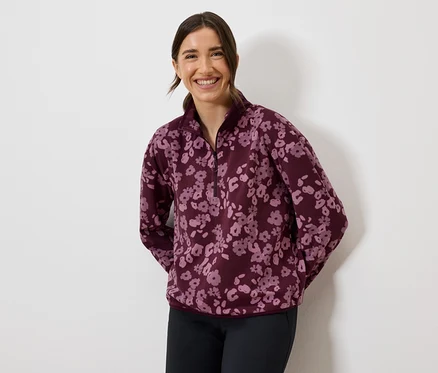Lächelnde Frau trägt ein burgunderfarbenes Fleece-Sweatshirt mit Blumenmuster und dunkle Hosen.