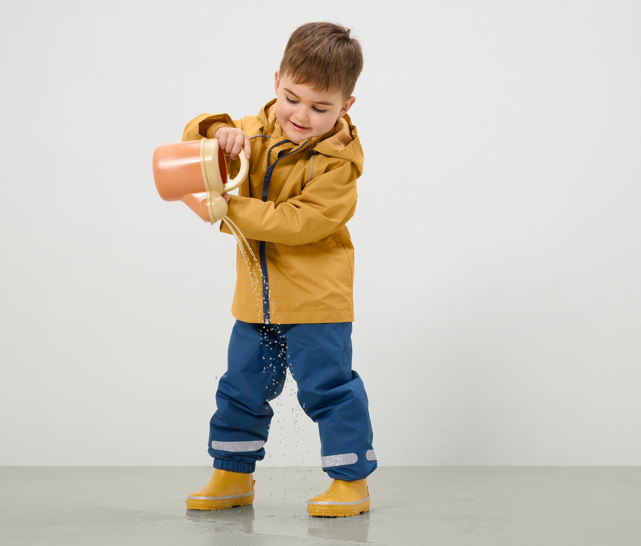 Junge in gelber Jacke und blauer Hose gießt Wasser aus einer orangefarbenen Gießkanne auf den Boden.