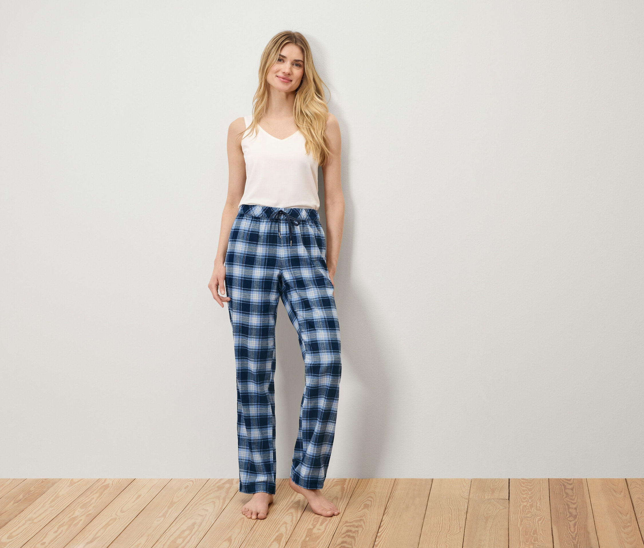 Eine Frau steht auf einem Holzboden und trägt ein weißes Tanktop und eine blau karierte Pyjamahose.