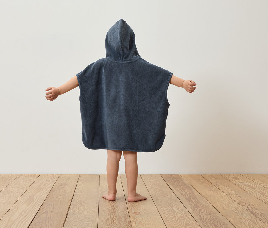 Kind steht mit dem Rücken zur Kamera und trägt einen marineblauen Poncho mit Kapuze und ausgestreckten Armen.