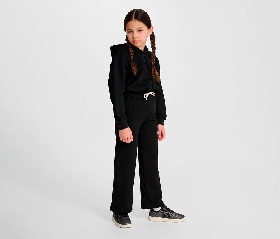 Mädchen mit Zöpfen steht vor einem Hintergrund und trägt einen schwarzen Hoodie und eine lockere HUMMEL HMLJR Loose WL Jogginghose.