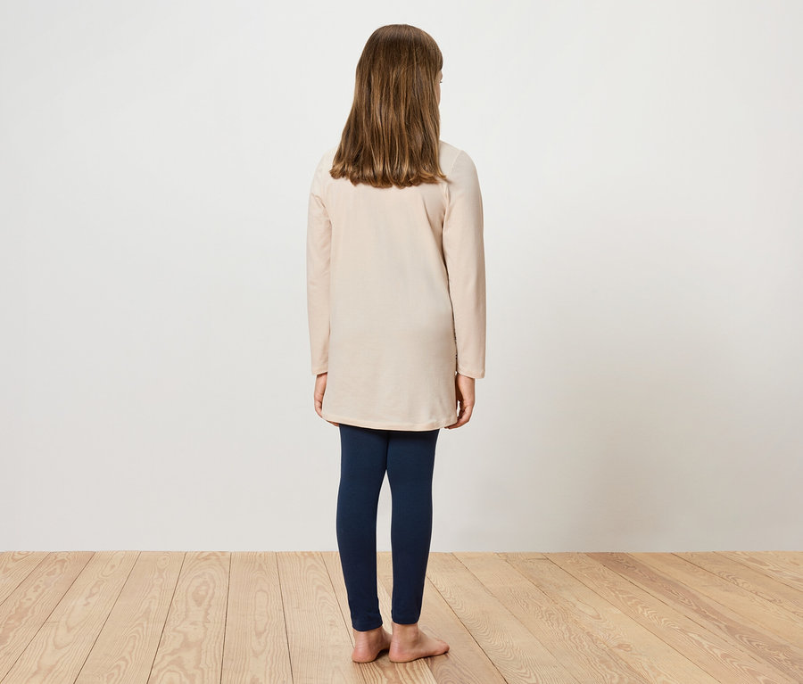 Mädchen steht mit dem Rücken zur Kamera, trägt eine cremefarbene Tunika und dunkelblaue Leggings.