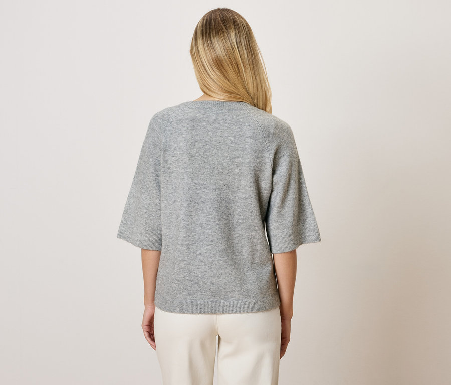 Blonde Frau von hinten in grauem Dreiviertelarm-Pullover und weißen Hosen.