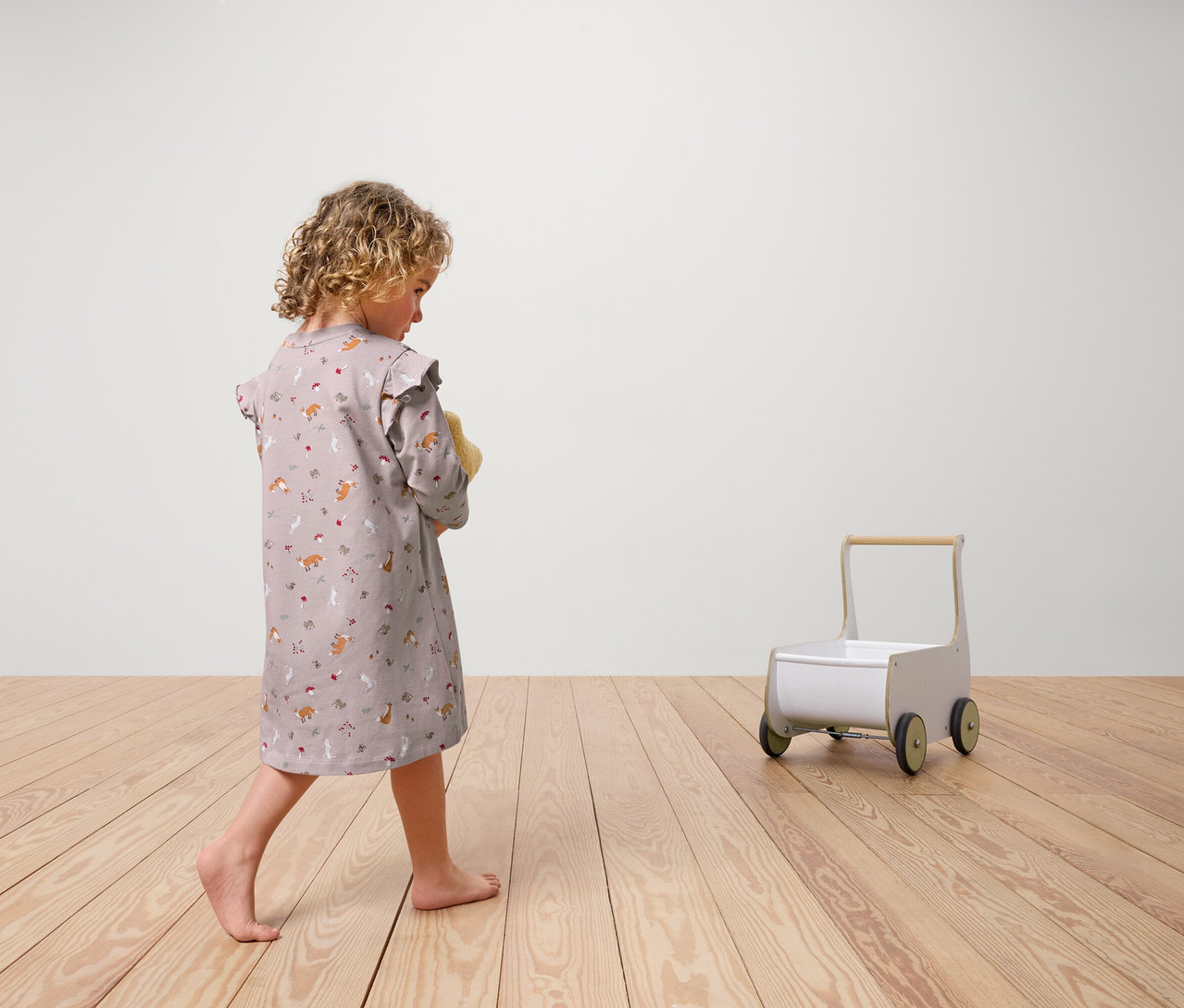 Mädchen von hinten, das ein 2 Kinder-Nachthemd trägt, hält ein Stofftier. Ein Spielzeug-Rollwagen steht auf einem Holzboden.