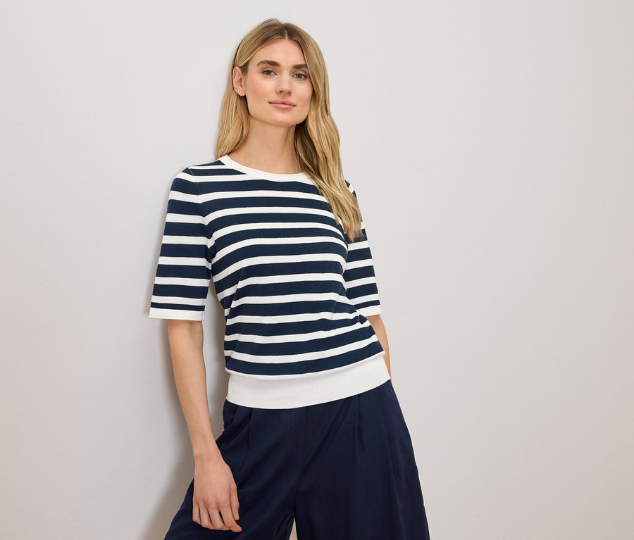 Frau mit blonden Haaren, die ein blau-weiß gestreiftes Oberteil und eine dunkelblaue Hose trägt.