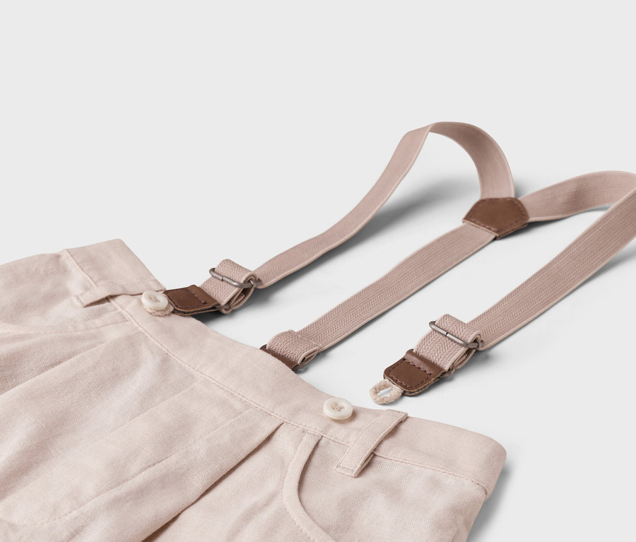 Nahaufnahme einer beigen Stoffhose mit Hosenträgern auf einem weißen Hintergrund.