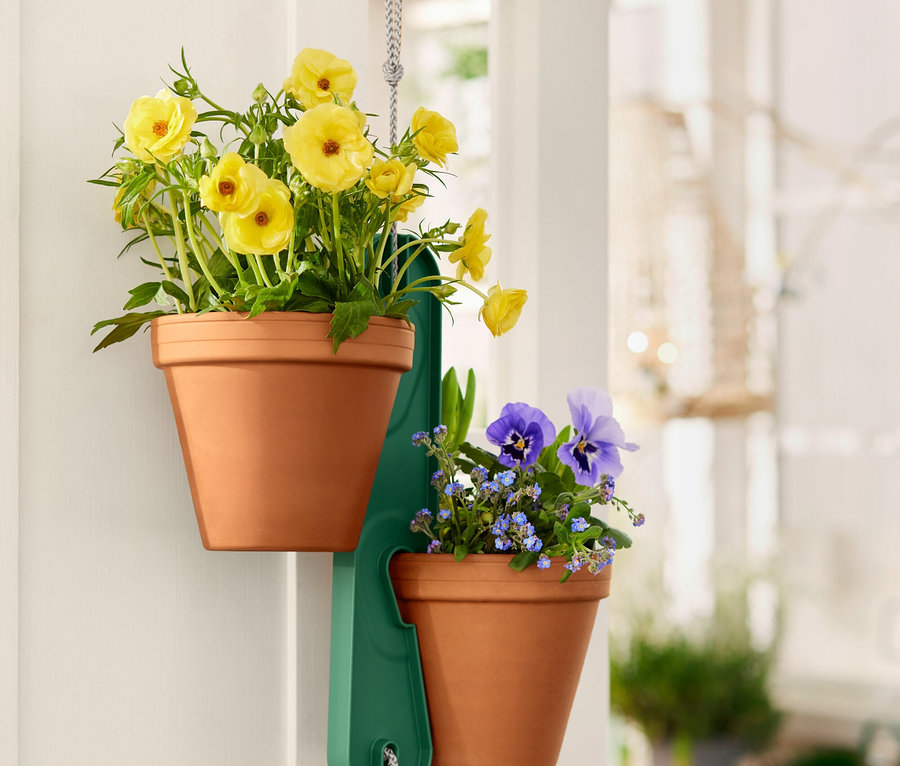 Zwei Terrakotta-Töpfe mit gelben bzw. blauen Blumen hängen an einem grünen Halter an einer weißen Tür.
