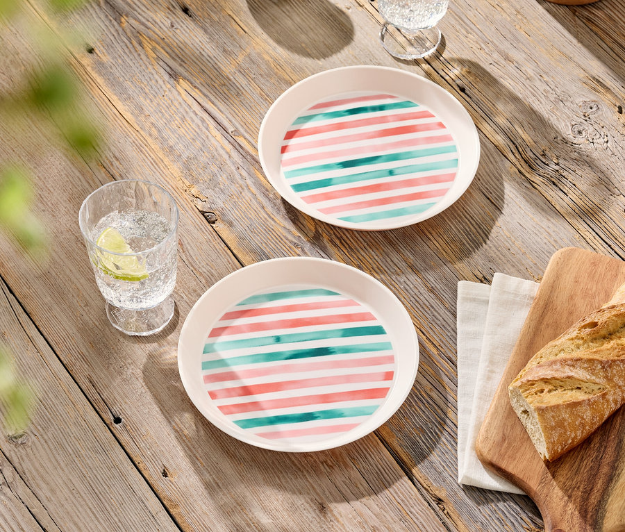 Zwei Teller mit rosa und türkisfarbenen Streifen, zwei Gläser mit Getränken, Oliven in einer Schale und Brot auf einem Schneidebrett auf einem Holztisch.