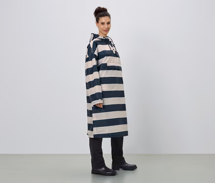 Frau steht seitlich, trägt einen langen, gestreiften Regenponcho mit Kapuze, schwarze Hose und Gummistiefel.