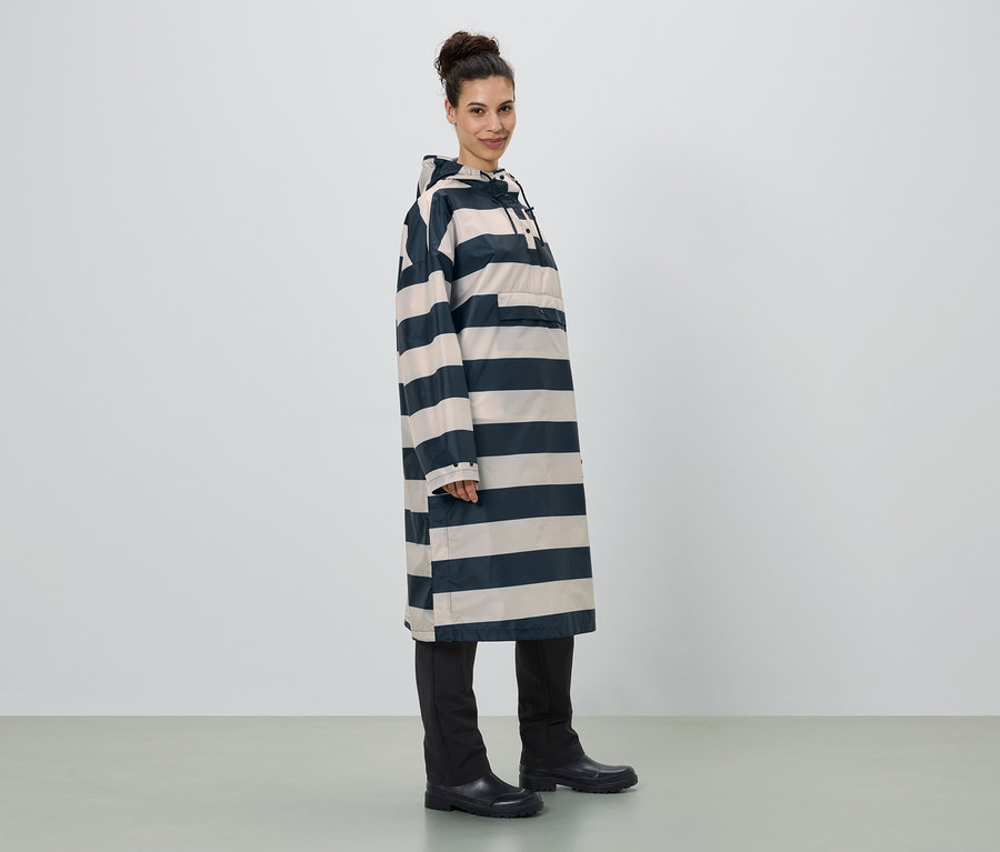 Frau steht seitlich, trägt einen langen, gestreiften Regenponcho mit Kapuze, schwarze Hose und Gummistiefel.