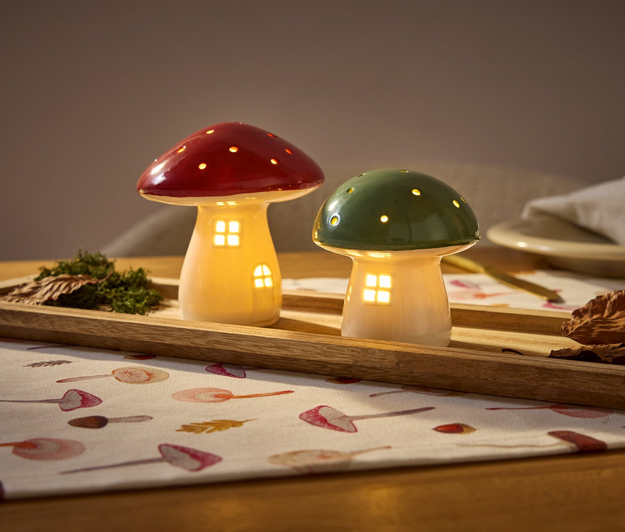 Zwei leuchtende Pilzhäuschen, eines rot und eines grün, stehen auf einem Holztablett mit Moos und Blättern auf einer Tischdecke.