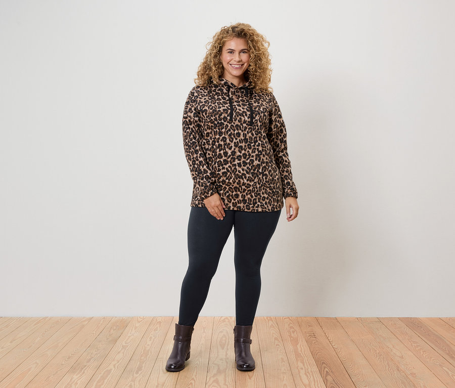 Eine Frau mit lockigem Haar steht und trägt ein Leopard-gemustertes Oberteil, Leggings und Stiefeletten.