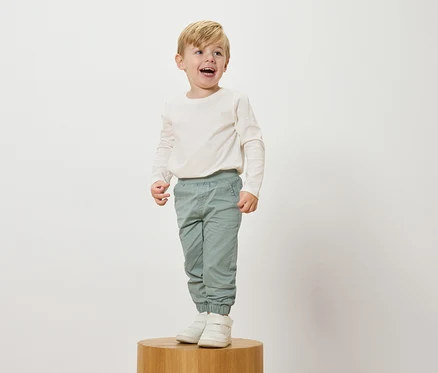 Ein lachender, blonder Junge steht auf einem Holzsockel, trägt ein langärmeliges, cremefarbenes Oberteil, hellblaue Hosen und weiße Turnschuhe.