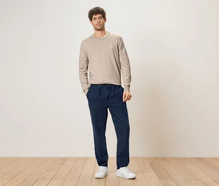 Ein Mann mit lockigem Haar steht mit den Händen in den Taschen vor einer Wand, er trägt einen hellbraunen Pullover, eine dunkelblaue Hose mit Kordelzug und weiße Turnschuhe.