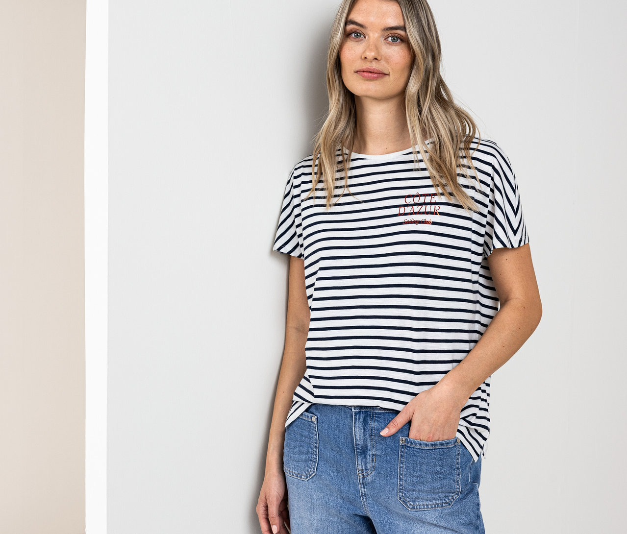 Frau mit blonden Haaren, die ein navy-weißes Street One T-Shirt und blaue Jeans trägt und an einer Wand lehnt.