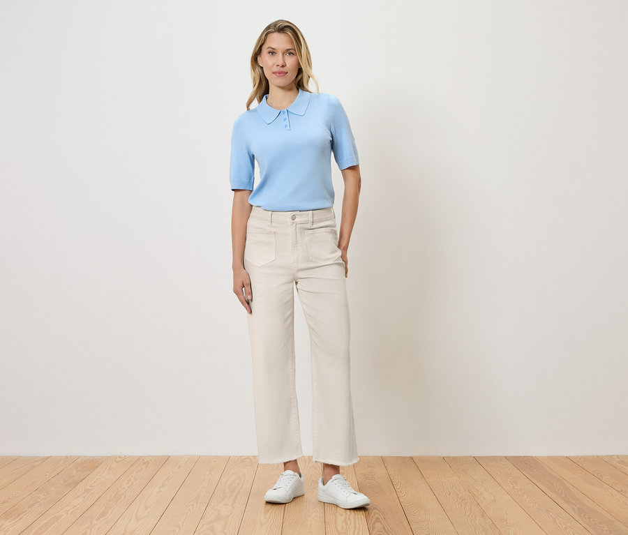 Frau steht vor einer Wand und trägt hellblaues Poloshirt, weiße Hose und weiße Schuhe.