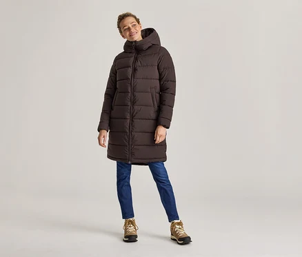 Frau in braunem, langem Puffer-Mantel, Jeans und Wanderschuhen steht vor hellem Hintergrund.