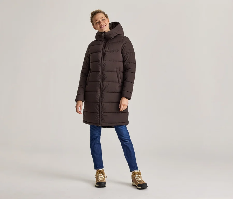 Frau in braunem, langem Puffer-Mantel, Jeans und Wanderschuhen steht vor hellem Hintergrund.
