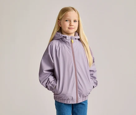 Mädchen mit blonden Haaren, in Jeans und lila Kapuzenjacke, die ihre Hände in den Taschen hat.
