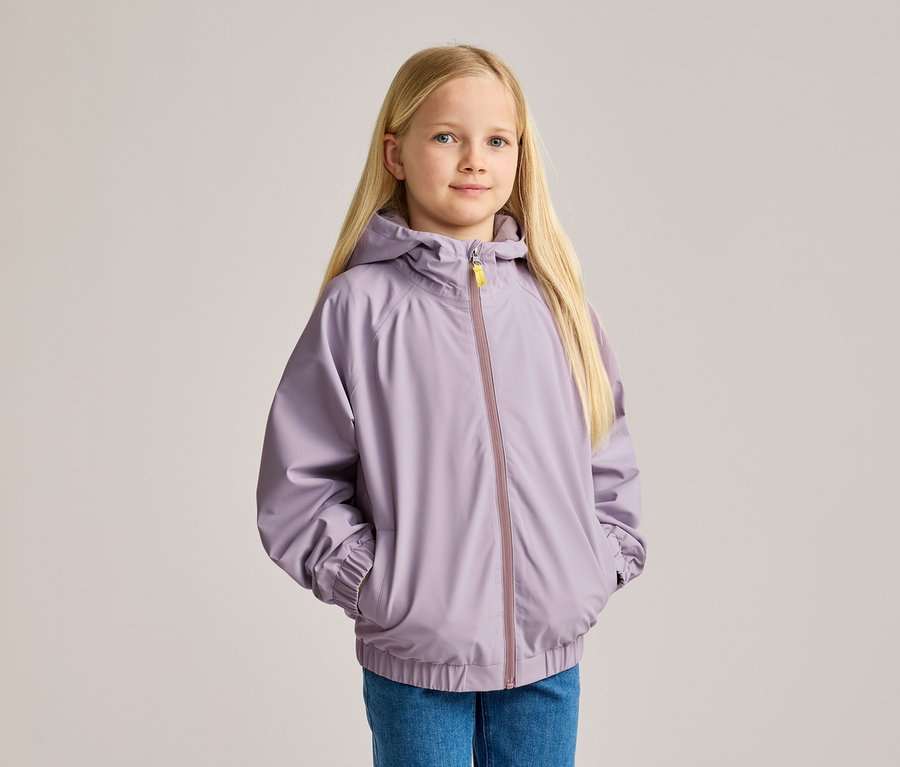Mädchen mit blonden Haaren, in Jeans und lila Kapuzenjacke, die ihre Hände in den Taschen hat.