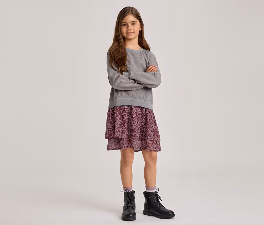 Ein junges Mädchen mit langen braunen Haaren steht mit verschränkten Armen und lächelt. Sie trägt ein graues Langarm-Sweatshirt, einen lila geblümten Stufenrock und schwarze Schnürstiefel mit hellvioletten Socken.