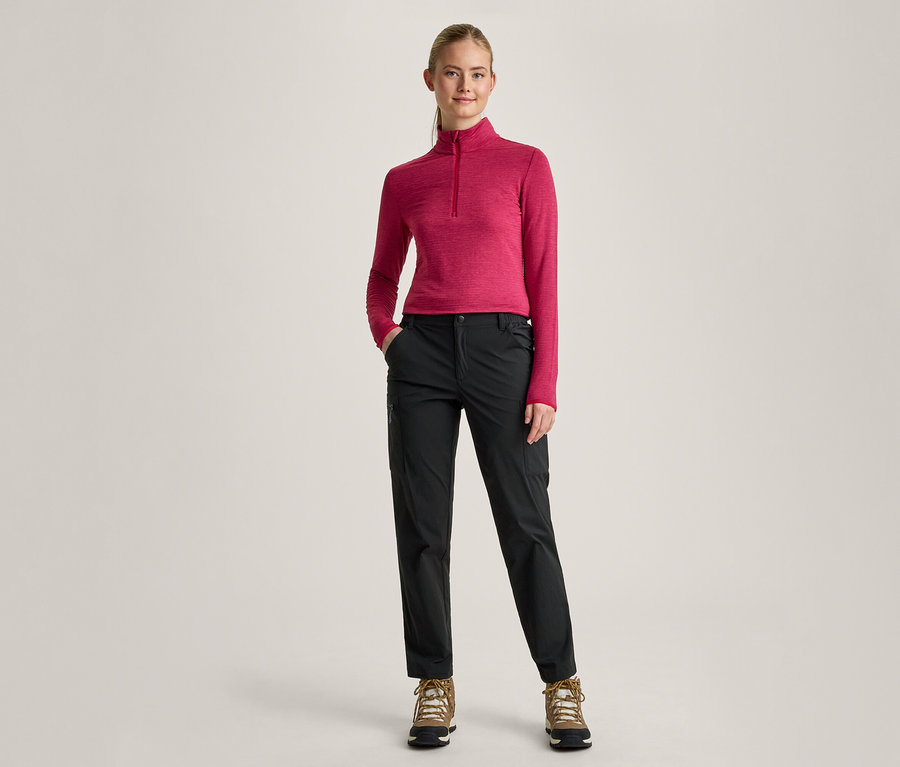Eine Frau mit blondem Pferdeschwanz steht mit den Händen in den Taschen nach vorne gerichtet. Sie trägt ein pinkes Langarmshirt mit Reißverschluss am Hals, schwarze Hosen und braune Wanderschuhe.