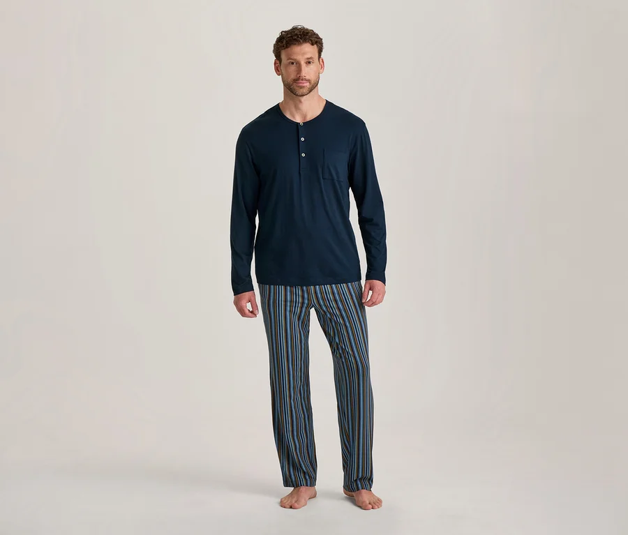 Mann steht barfuß, trägt ein dunkelblaues, langärmeliges Pyjama-Oberteil mit Knöpfen und Tasche sowie gestreifte Pyjamahosen in Blau, Braun und Schwarz.