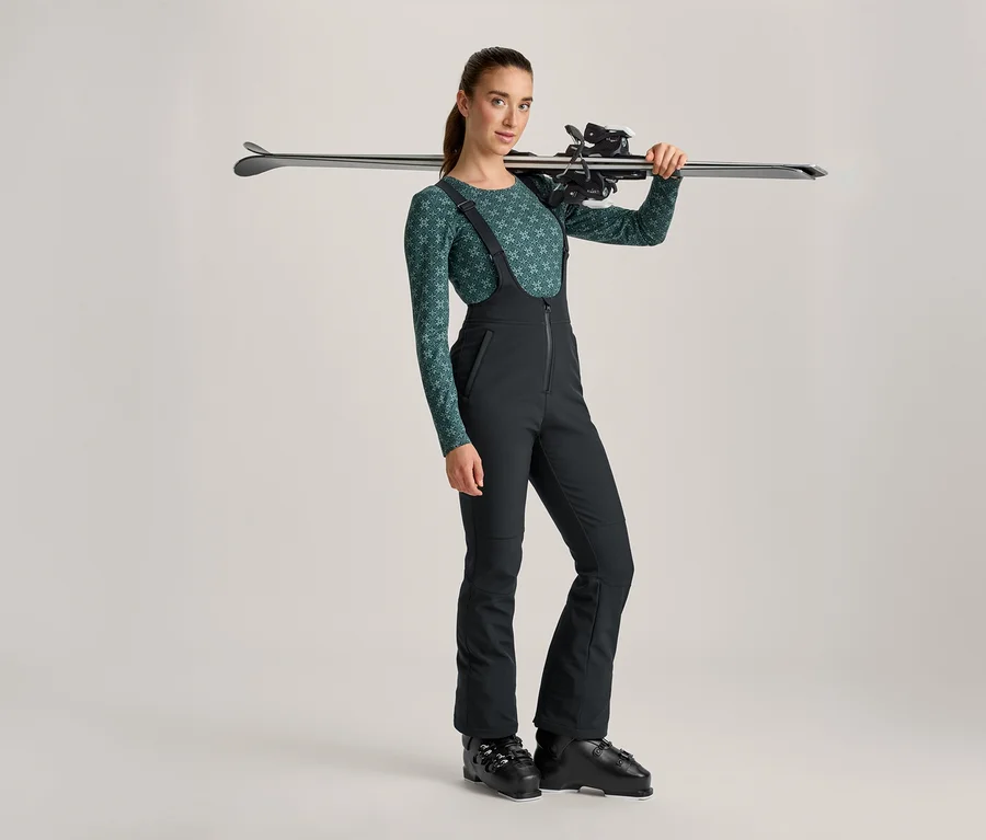 Frau in grün gemustertem Langarmshirt und schwarzer Skihose mit Skischuhen, Skier über der Schulter tragend.