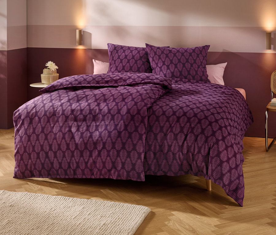 Ein Bett mit violetter Bettwäsche mit Blattmuster steht in einem Schlafzimmer mit zweifarbigen Wänden, Holzboden und einem hellen Teppich. Zwei Wandlampen beleuchten den Bereich über dem Bett. Links steht ein kleiner Beistelltisch mit einer Blume, rechts ein Stuhl mit Büchern.