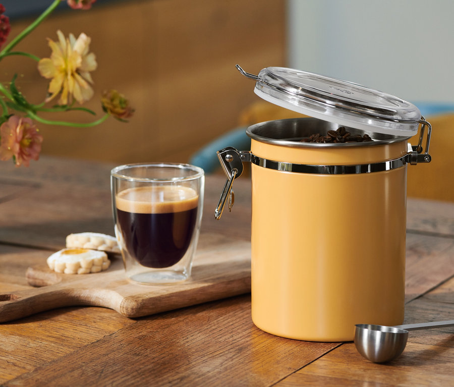 Kaffee in einem Glas, Kekse auf einem Holzbrett und ein offener gelber Kaffeebehälter mit Bohnen, dazu ein Messlöffel, auf einem Holztisch mit Blumen.